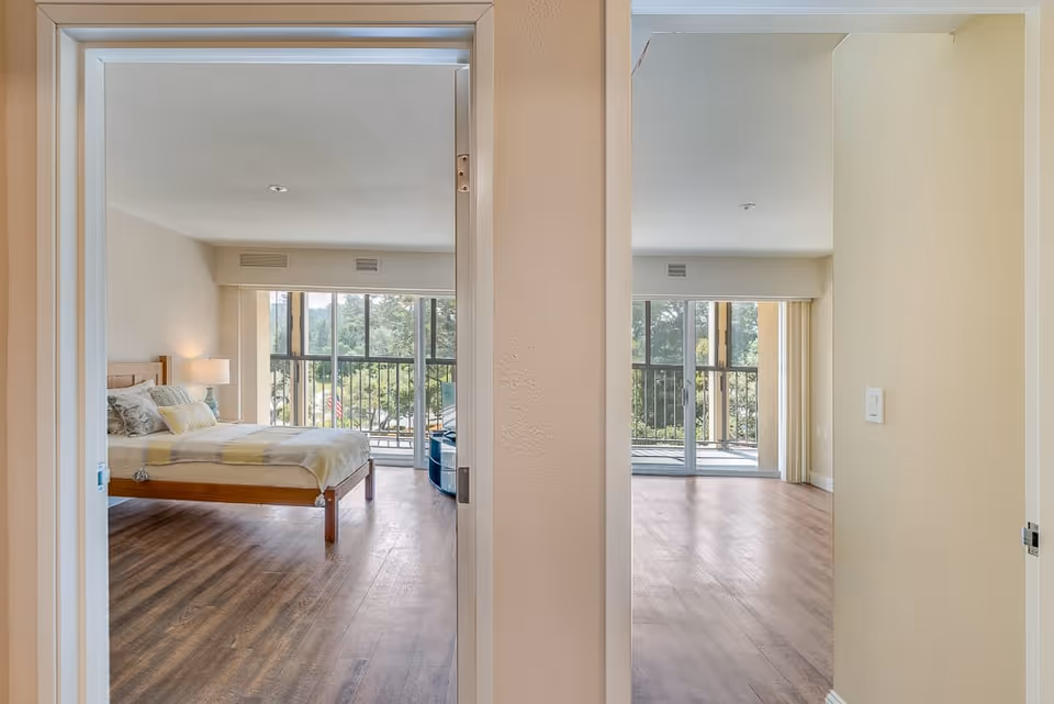 View through two adjacent doorways showing a bedroom on the left with a bed, lamp, and large windows overlooking a balcony with trees outside, and an empty room on the right with large windows and a sliding glass door leading to a balcony.