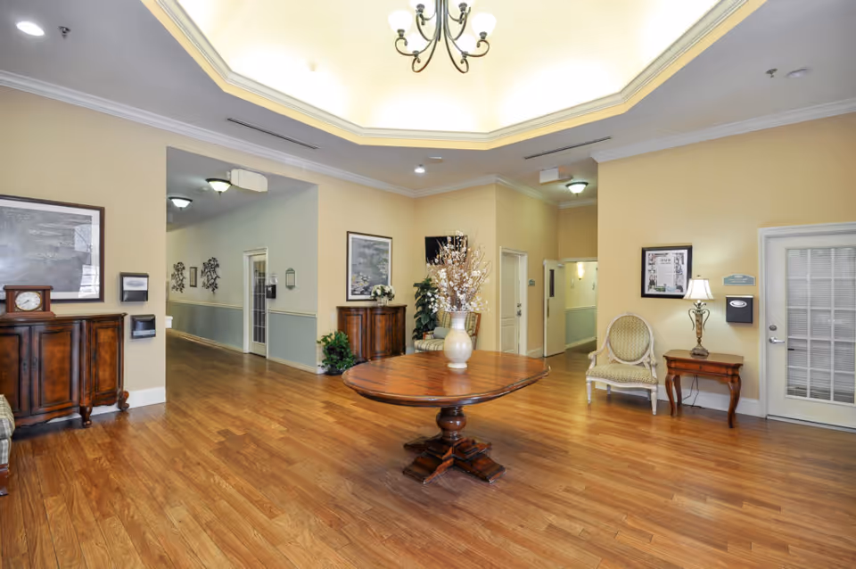 A spacious and well-lit interior common area with wooden flooring, beige walls, and a decorative ceiling light fixture. The room features a round wooden table with a vase of flowers in the center, two wooden cabinets with framed artwork above them, a cushioned chair, a small side table with a lamp, and a hallway leading to other rooms.