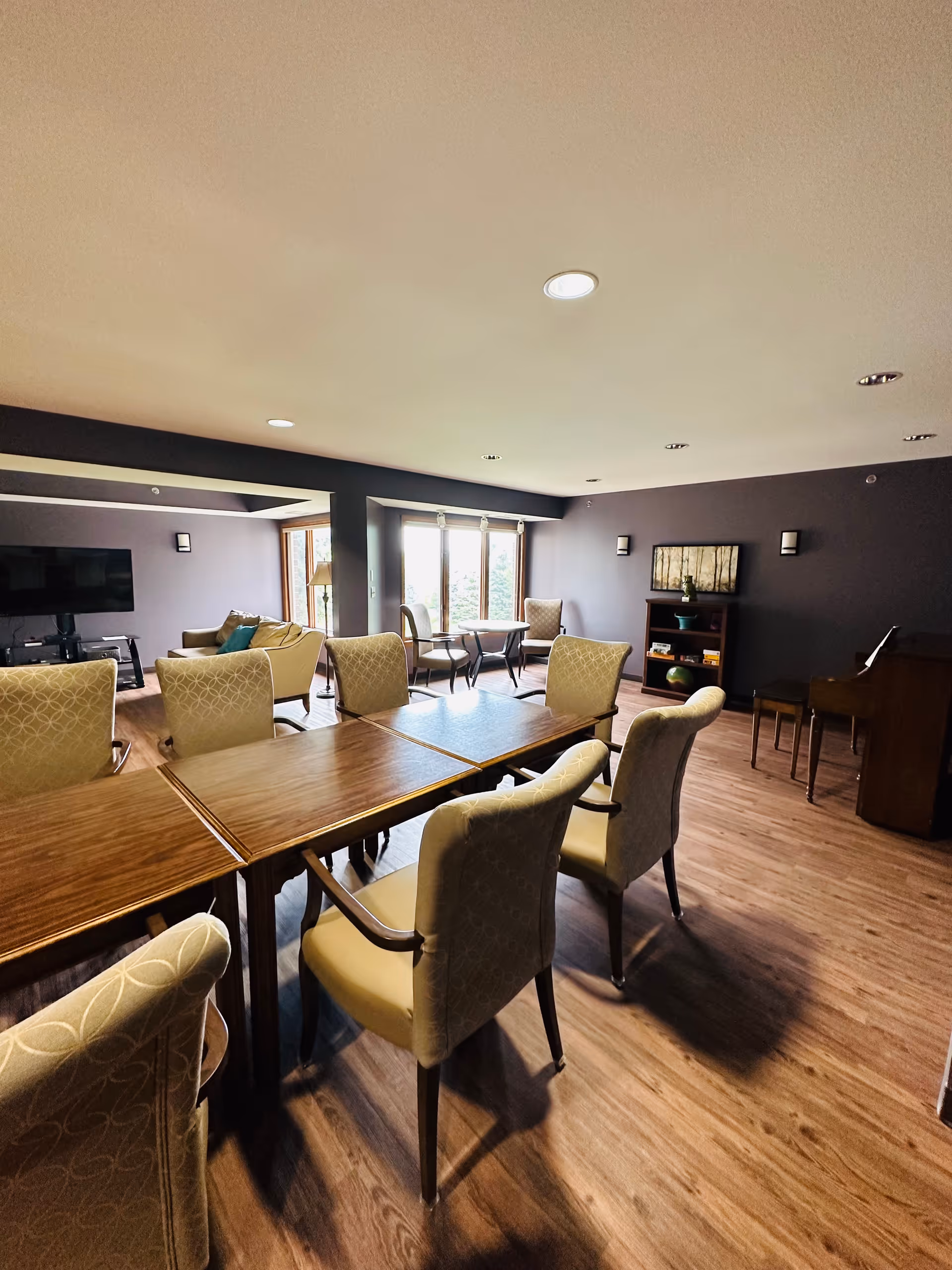 A spacious senior living common area with several upholstered chairs arranged around wooden tables. The room has wood flooring, a dark accent wall with a bookshelf and framed artwork, a piano with a bench, and a large flat-screen TV mounted on the wall. Large windows allow natural light to enter the room.