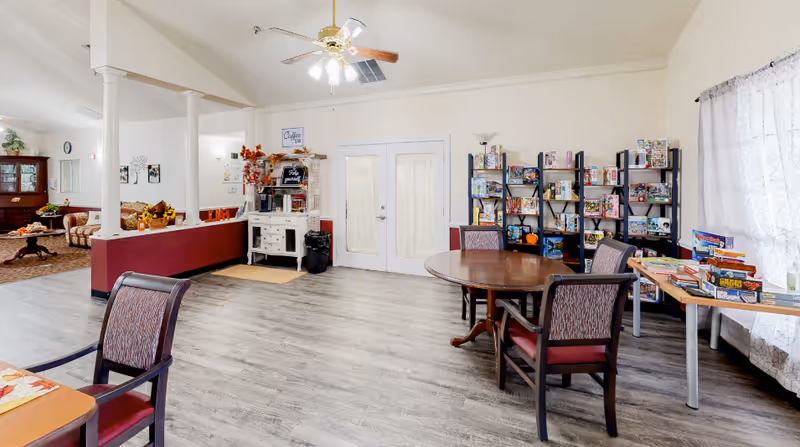 Bright communal activity room with tables and chairs, bookshelves of games and puzzles, a coffee station, and double doors.