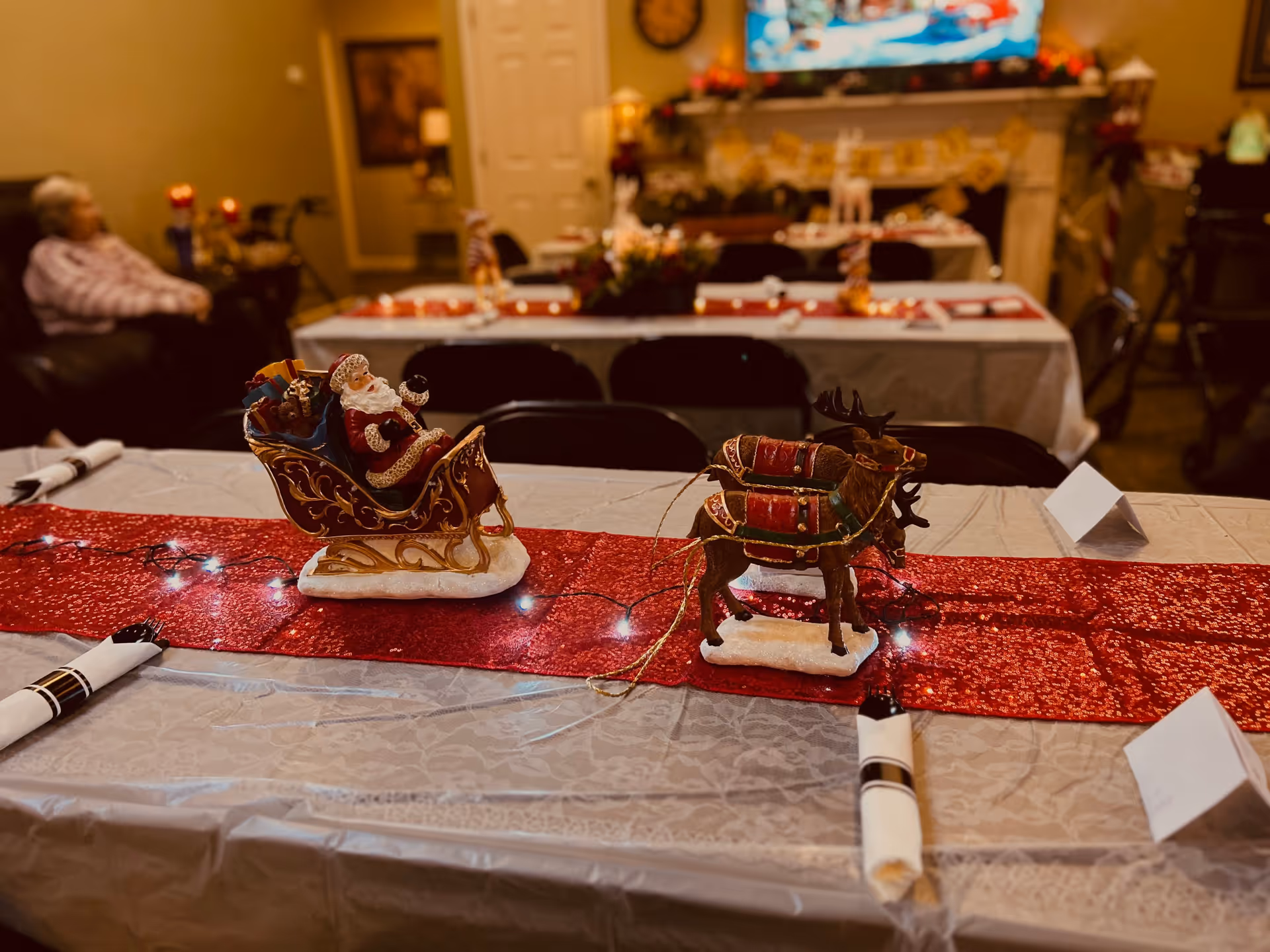 Festively decorated dining table with a Santa sleigh and reindeer centerpiece, red runner, and place settings in a communal dining room.