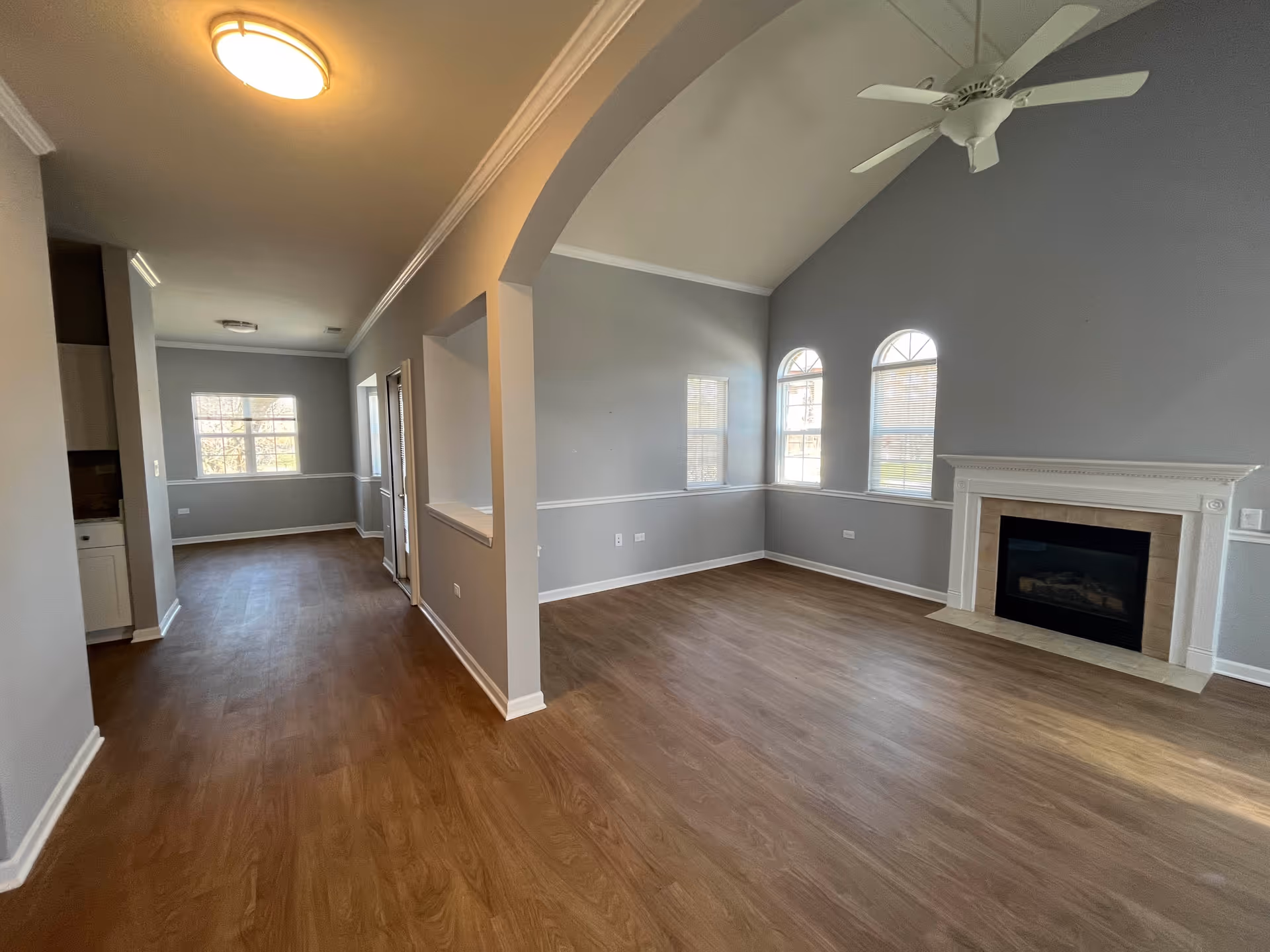 An empty interior living space with light gray walls, wood flooring, a white ceiling fan, and a fireplace with a white mantel. The room has three windows with blinds, two of which are arched. There is an open archway leading to another room with a window at the far end.