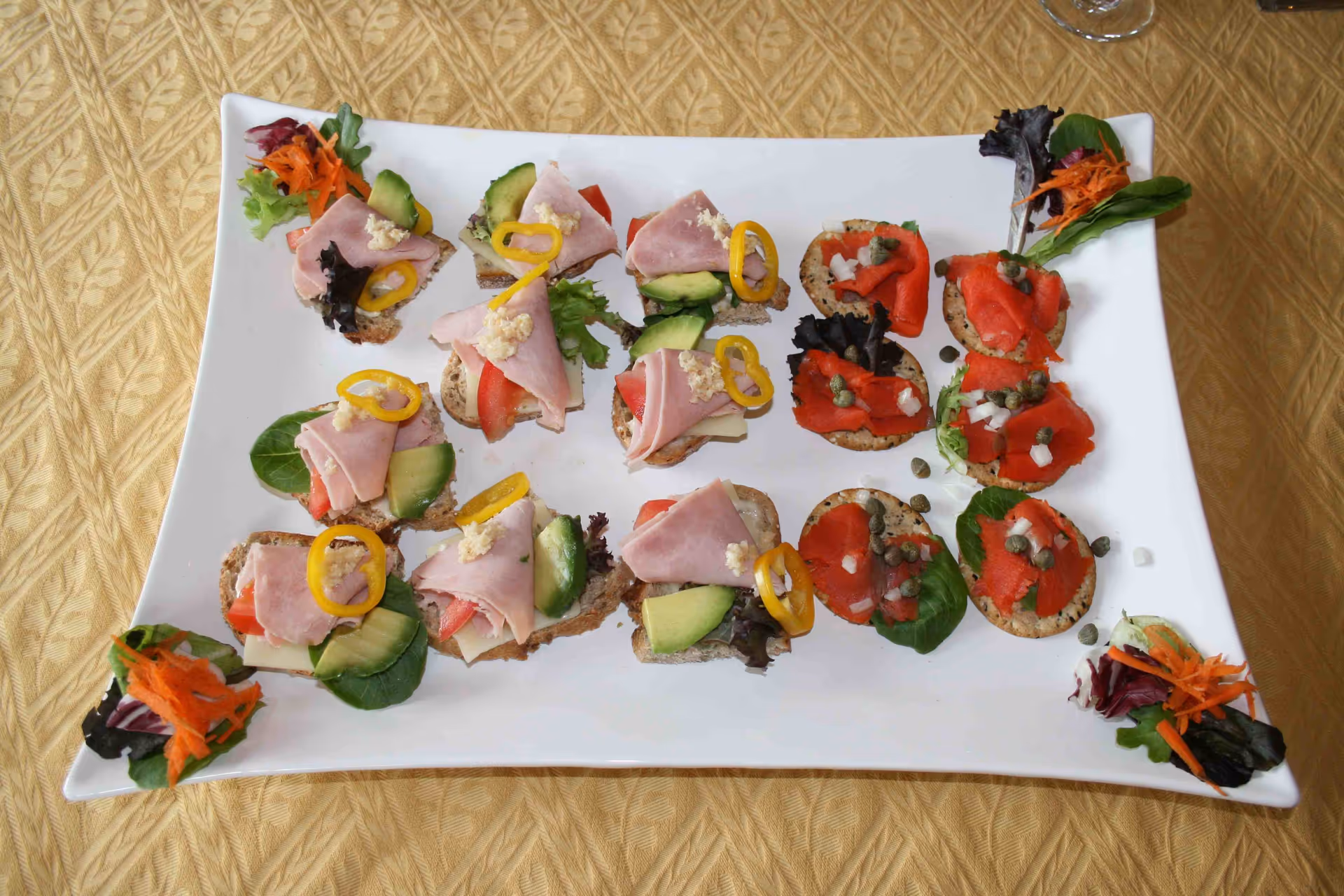 A white rectangular plate with assorted finger sandwiches and crackers topped with ham, avocado, tomato, yellow pepper rings, smoked salmon, capers, and garnished with leafy greens and shredded carrots on a gold patterned tablecloth.