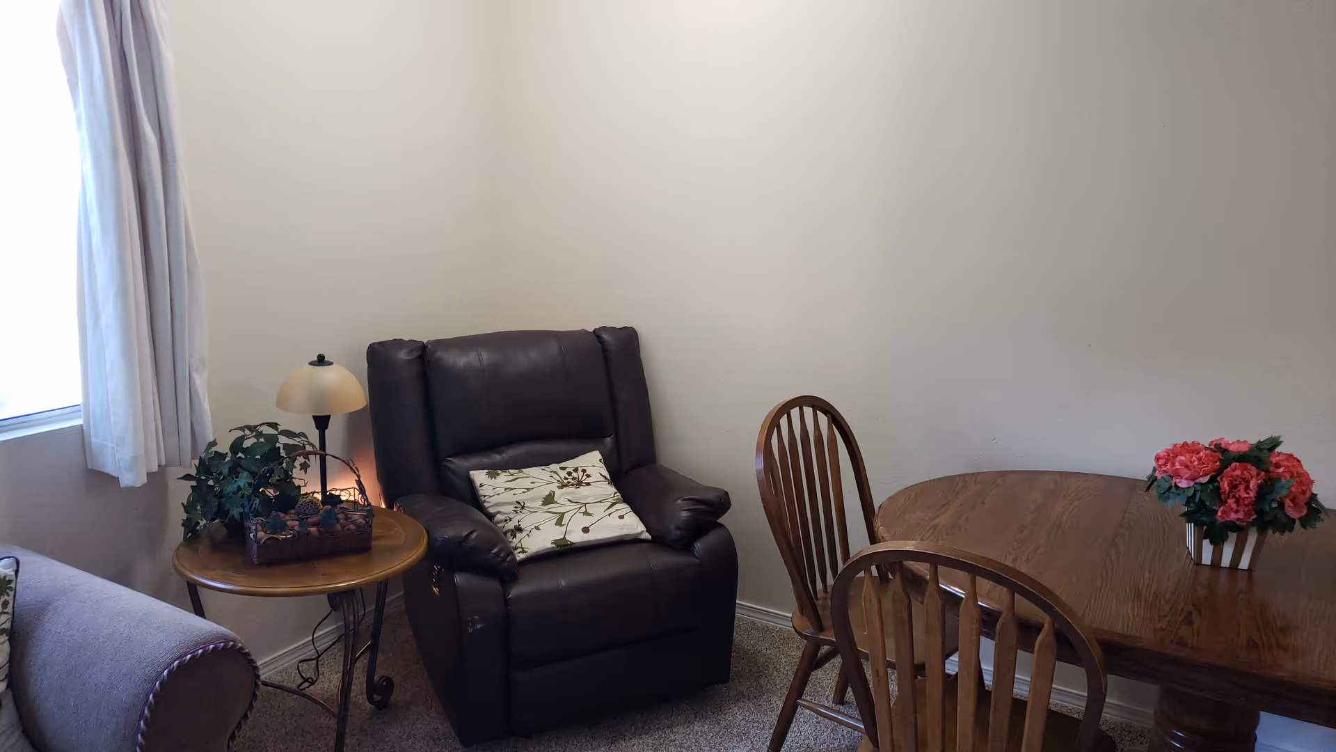 A cozy corner of a room featuring a dark brown leather recliner with a floral cushion, a small round wooden side table with a lamp and a basket of greenery, a wooden dining table with two wooden chairs, and a vase of pink flowers on the table. Light-colored walls and carpeted floor complete the setting.