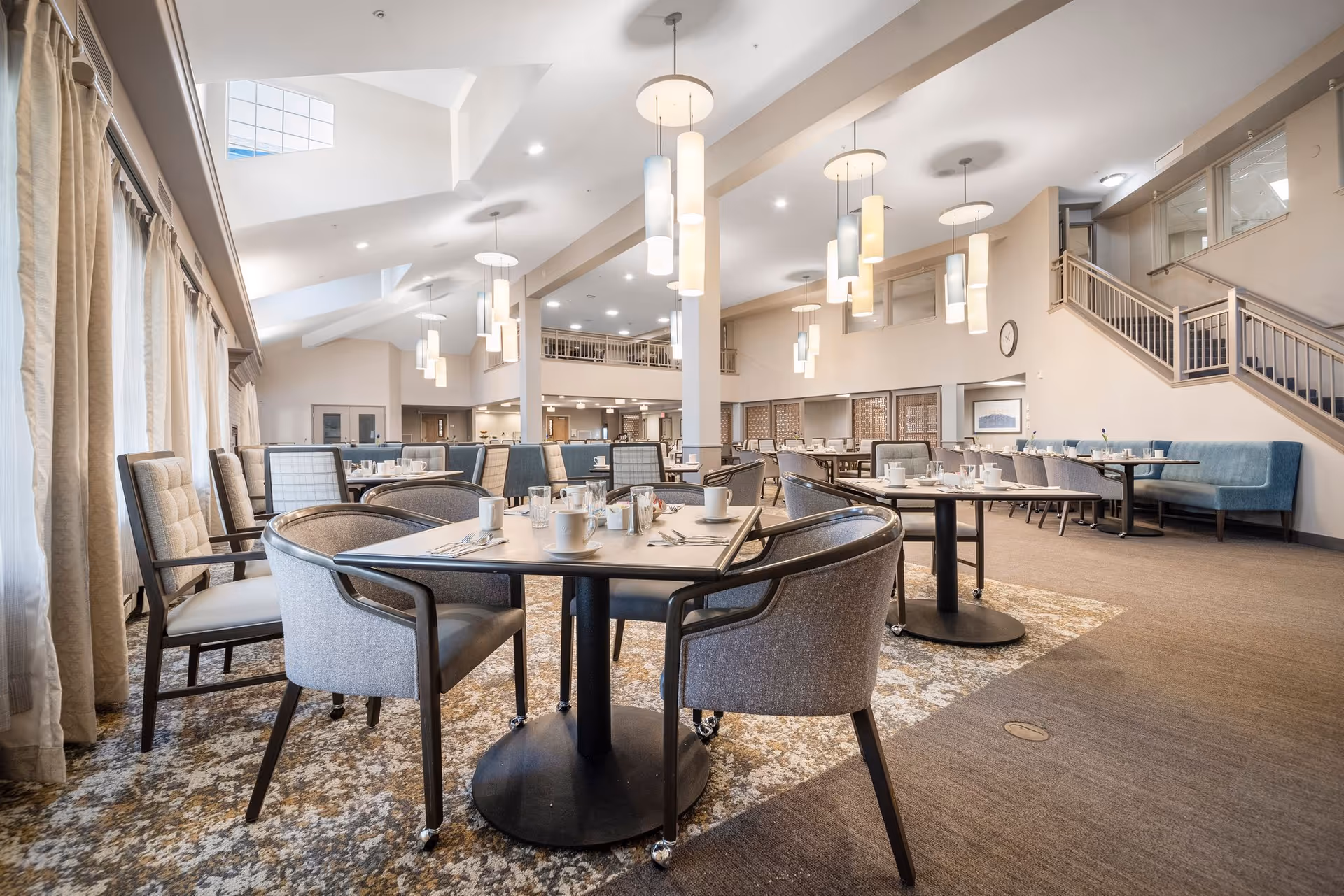 Spacious bright dining room with round tables and upholstered chairs set for meals under hanging pendant lights.