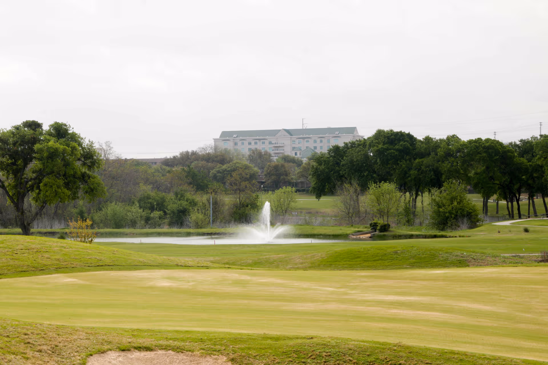 A scenic view of a golf course with a sand bunker in the foreground, a water fountain in a pond in the middle ground, and a large building partially obscured by trees in the background under a cloudy sky.