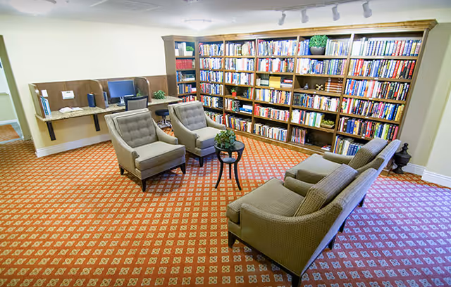 A cozy library room with a large wooden bookshelf filled with books along one wall. In front of the bookshelf, there are four cushioned armchairs arranged around a small round table with a plant on it. To the left, there is a small desk area with two computer monitors and chairs. The floor is covered with a patterned carpet, and the room is well-lit with ceiling lights.