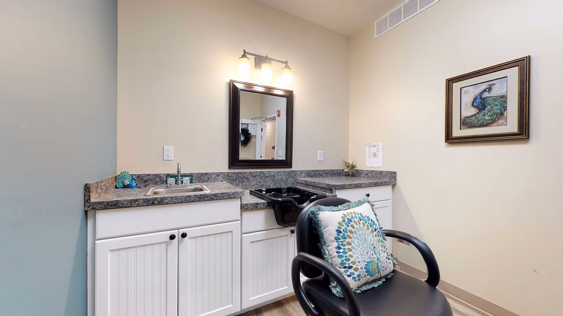 A small salon area in a retirement living facility featuring a black salon chair with a decorative pillow, a countertop with a sink, a black hair washing basin, a mirror with three lights above it, white cabinets below the countertop, and a framed peacock artwork on the wall.