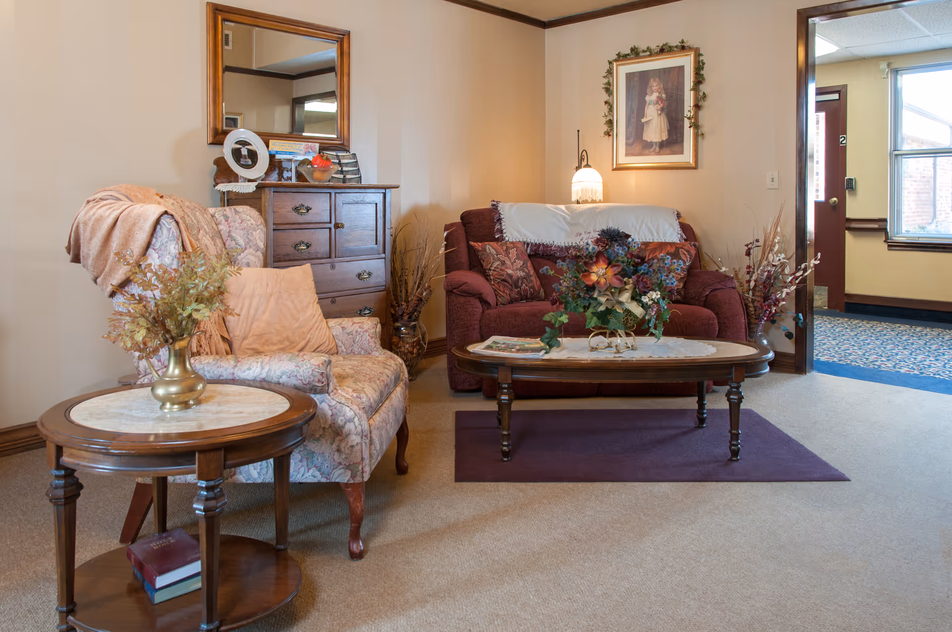 A cozy living room area with a floral upholstered armchair draped with a peach-colored blanket, a wooden side table with a vase of dried flowers, a wooden chest of drawers with a mirror above it, and a maroon loveseat with decorative pillows and a white throw. A wooden coffee table with a floral arrangement and magazines sits on a purple rug. The room has beige walls and carpet, with a framed picture hanging above the loveseat and a doorway leading to a hallway with a window.
