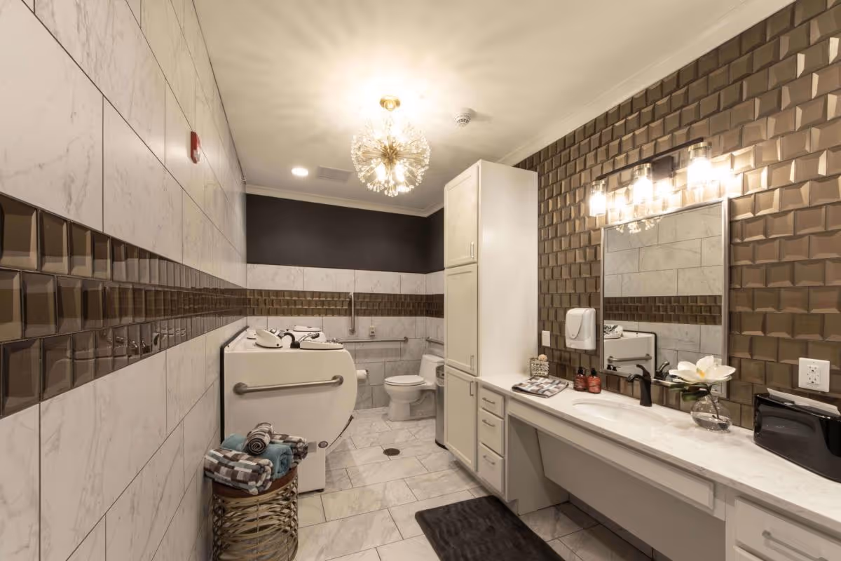 A modern, well-lit bathroom featuring a white toilet, a large white bathtub with safety rails, and a long vanity with a sink, mirror, and decorative lighting. The walls are decorated with white and brown tiles, and there is a basket with folded towels near the bathtub. A flower in a glass vase and soap dispensers are placed on the vanity countertop.