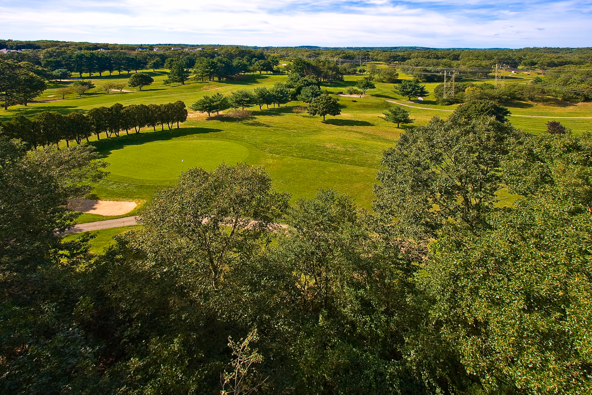 Aerial view of a lush green golf course with trees, fairways, and a sand bunker under a partly cloudy sky.