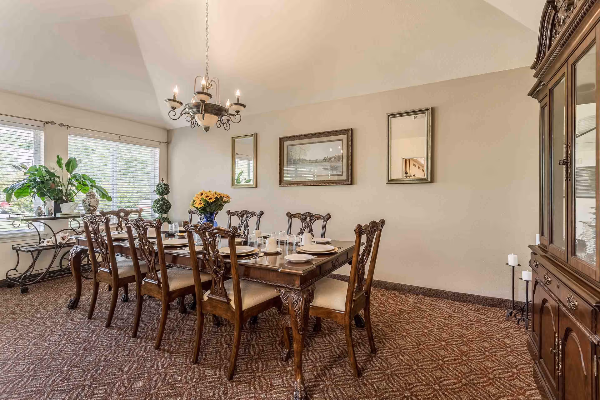 A formal dining room with a large wooden dining table set for eight people with plates, cups, and glasses. The room features a chandelier hanging from a vaulted ceiling, a large window with blinds, decorative plants, a side table, and a wooden china cabinet. The walls are adorned with two mirrors and a framed painting.