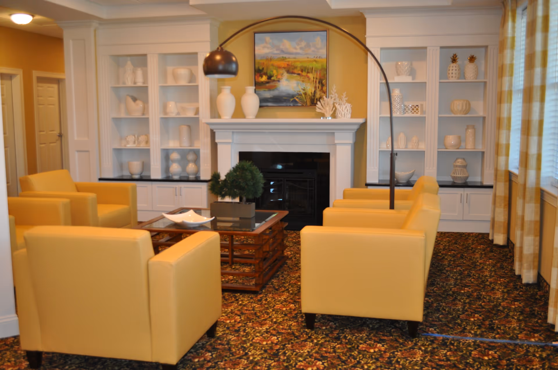 A cozy living room area with four yellow armchairs arranged around a wooden coffee table. The room features a white fireplace with decorative vases on the mantel and built-in white shelves filled with various white ceramic decor items. A large arc floor lamp stands behind one of the chairs, and a colorful landscape painting hangs above the fireplace. The room has patterned carpet and yellow checkered curtains on the windows.