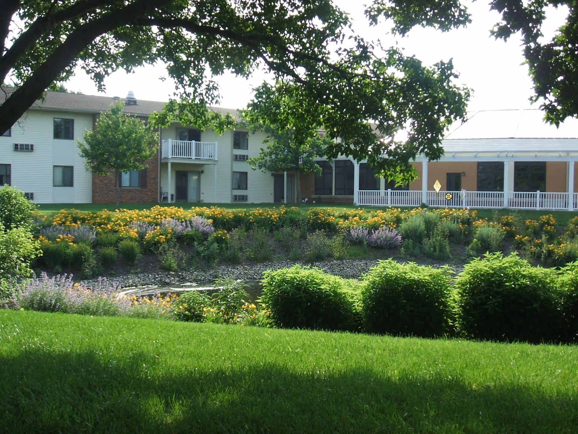 View of a retirement community building with a well-maintained garden in the foreground. The garden features green grass, bushes, and colorful flowers with a small pond or water feature. The building has white siding, brick accents, balconies, and a covered porch area. Trees frame the top of the image providing shade.