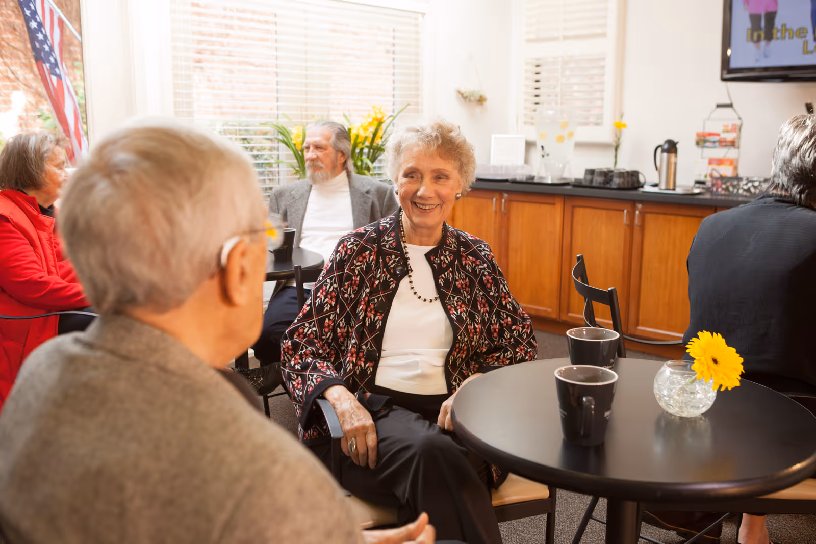 A group of elderly people sitting and chatting in a bright common area with large windows. There are small round tables with black chairs, black coffee mugs, and a small vase with a yellow flower on one table. The room has wooden cabinets along one wall and a television mounted above them.