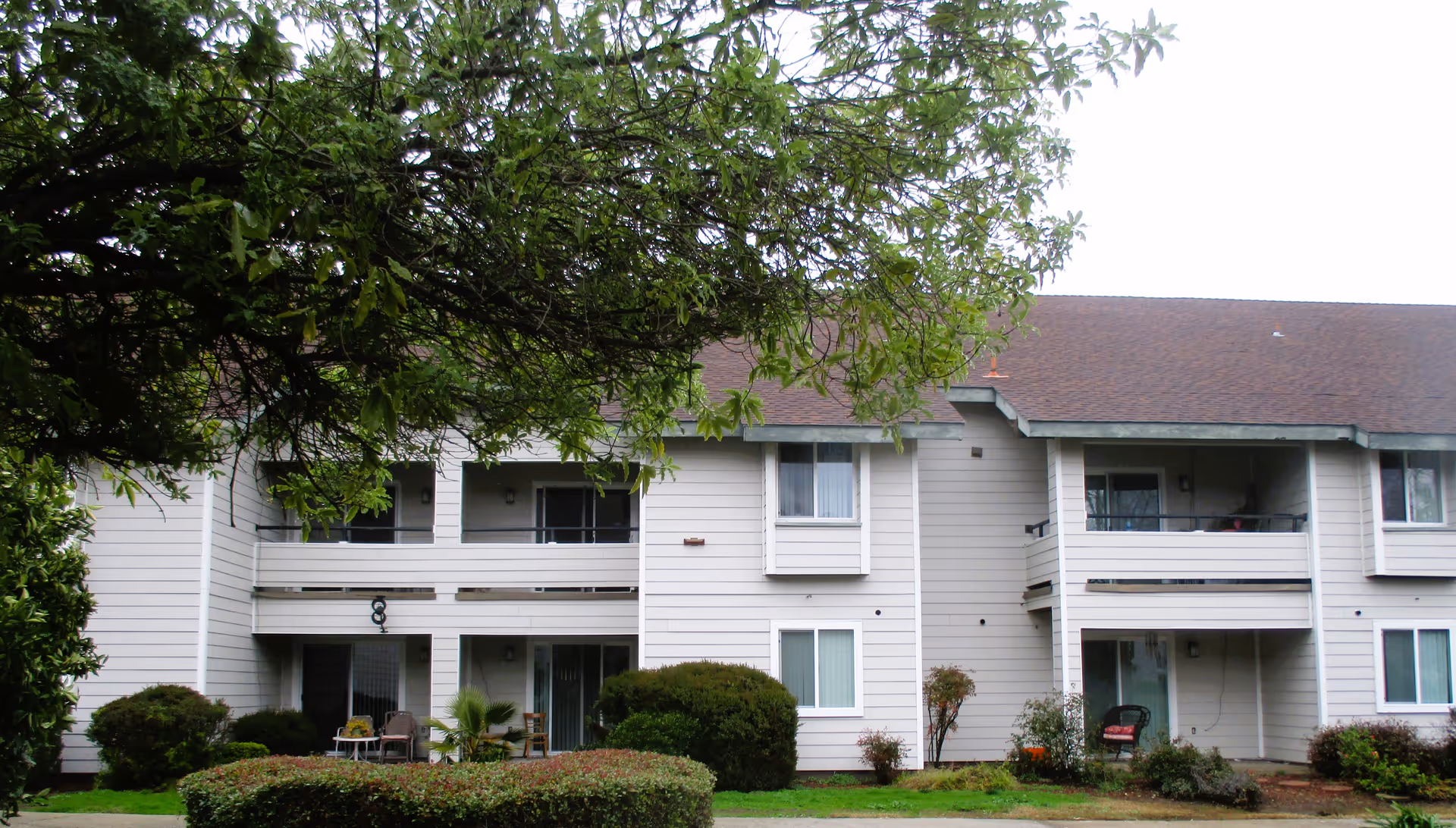 Exterior view of a two-story residential building with balconies and patios, surrounded by green bushes and trees under an overcast sky.