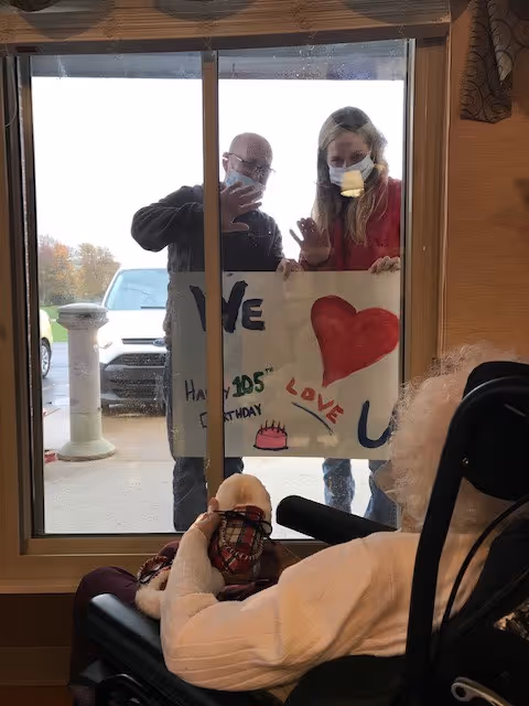 An elderly person sitting in a wheelchair inside a room, looking out through a glass door where two people wearing face masks stand outside holding a handmade sign that says 'We love U' with a red heart and a birthday cake drawing. The people outside are waving to the elderly person.