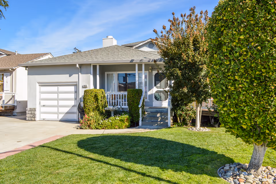 Front exterior view of a single-story house with a small porch, white garage door, trimmed bushes, green lawn, and trees under a clear blue sky.