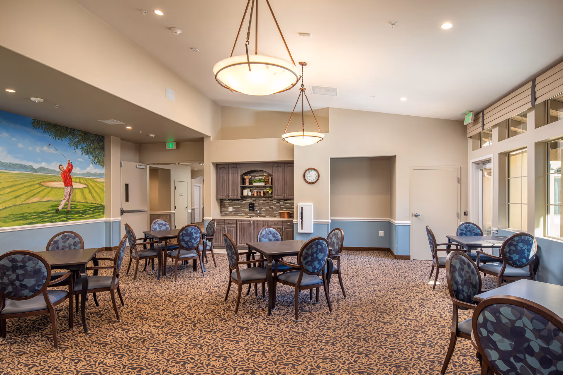 A spacious dining area with multiple tables and chairs featuring blue patterned upholstery. The room has beige walls with a blue wainscot, carpeted floor with an intricate pattern, and two large hanging light fixtures. There is a kitchenette with cabinets, a clock on the wall, and a mural of a golfer swinging a club on one side. Large windows allow natural light to enter the room.