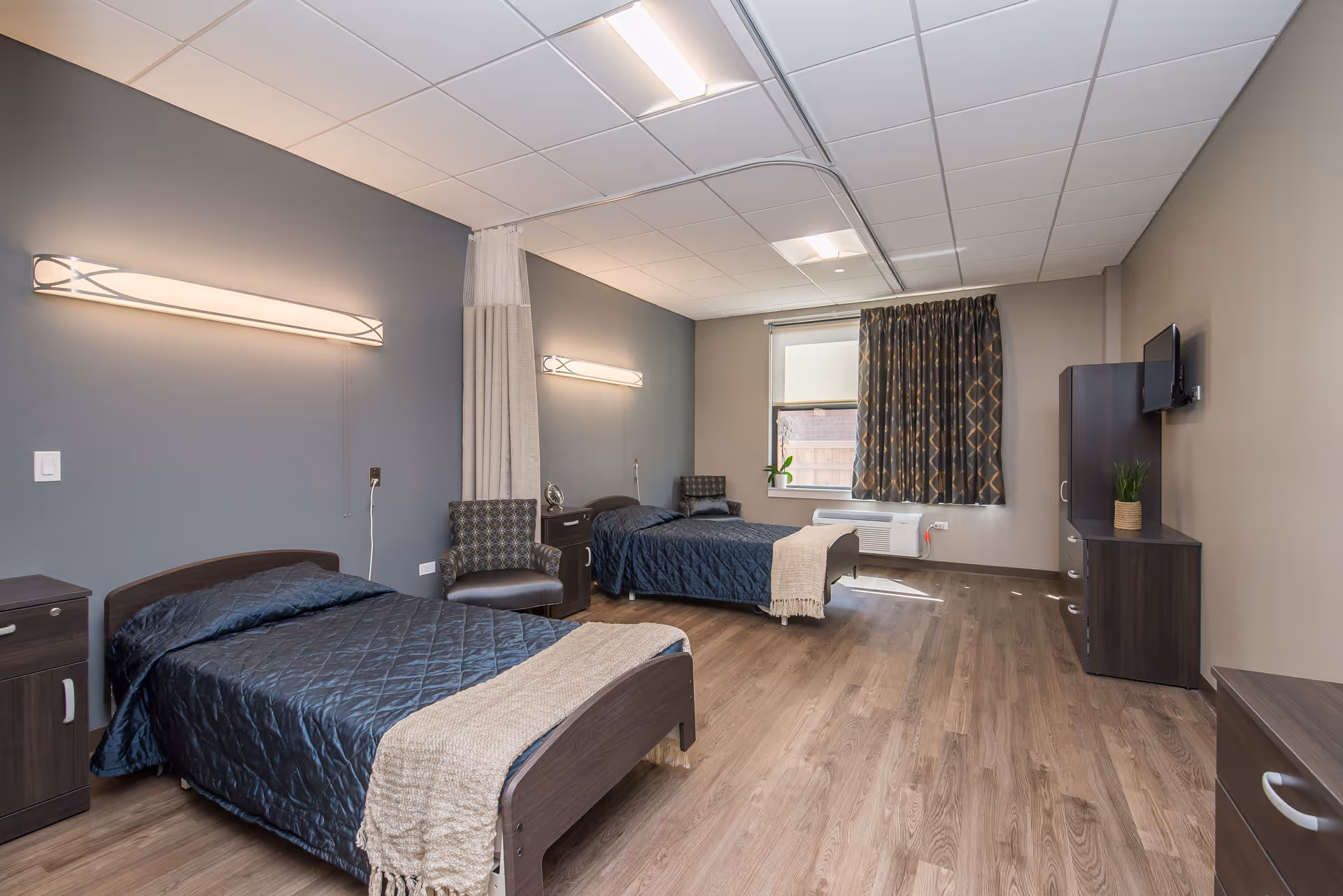 A spacious bedroom with two single beds, each covered with dark blue quilts and beige throws. The room features wood flooring, a window with patterned curtains, a wall-mounted TV, a dark wood dresser, and two chairs. The walls are painted in neutral tones with modern light fixtures above each bed.