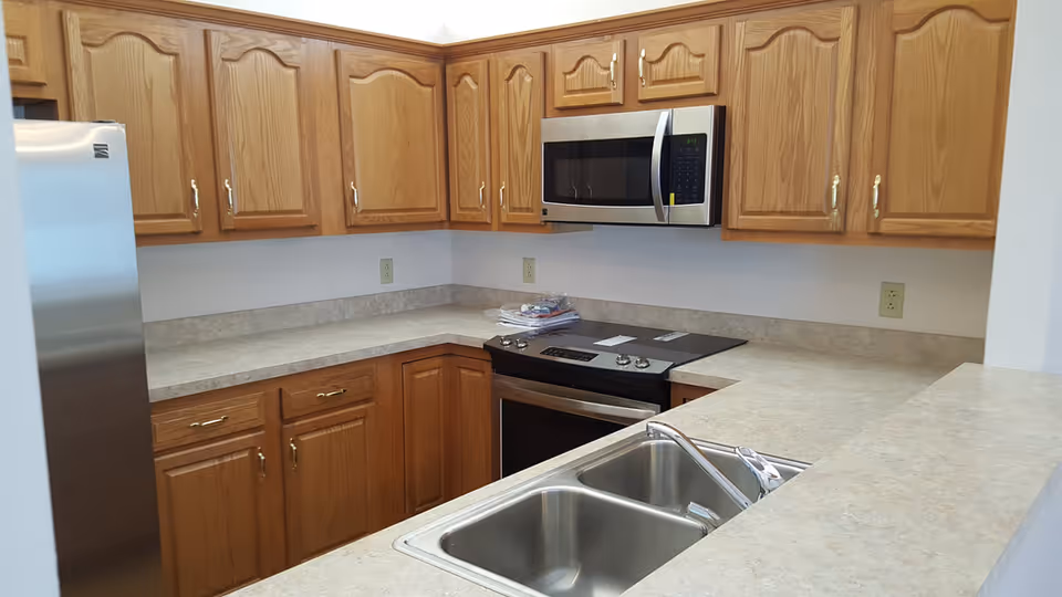 A kitchen with wooden cabinets, a stainless steel refrigerator, a built-in microwave above an electric stove, and a double stainless steel sink set in a beige countertop.