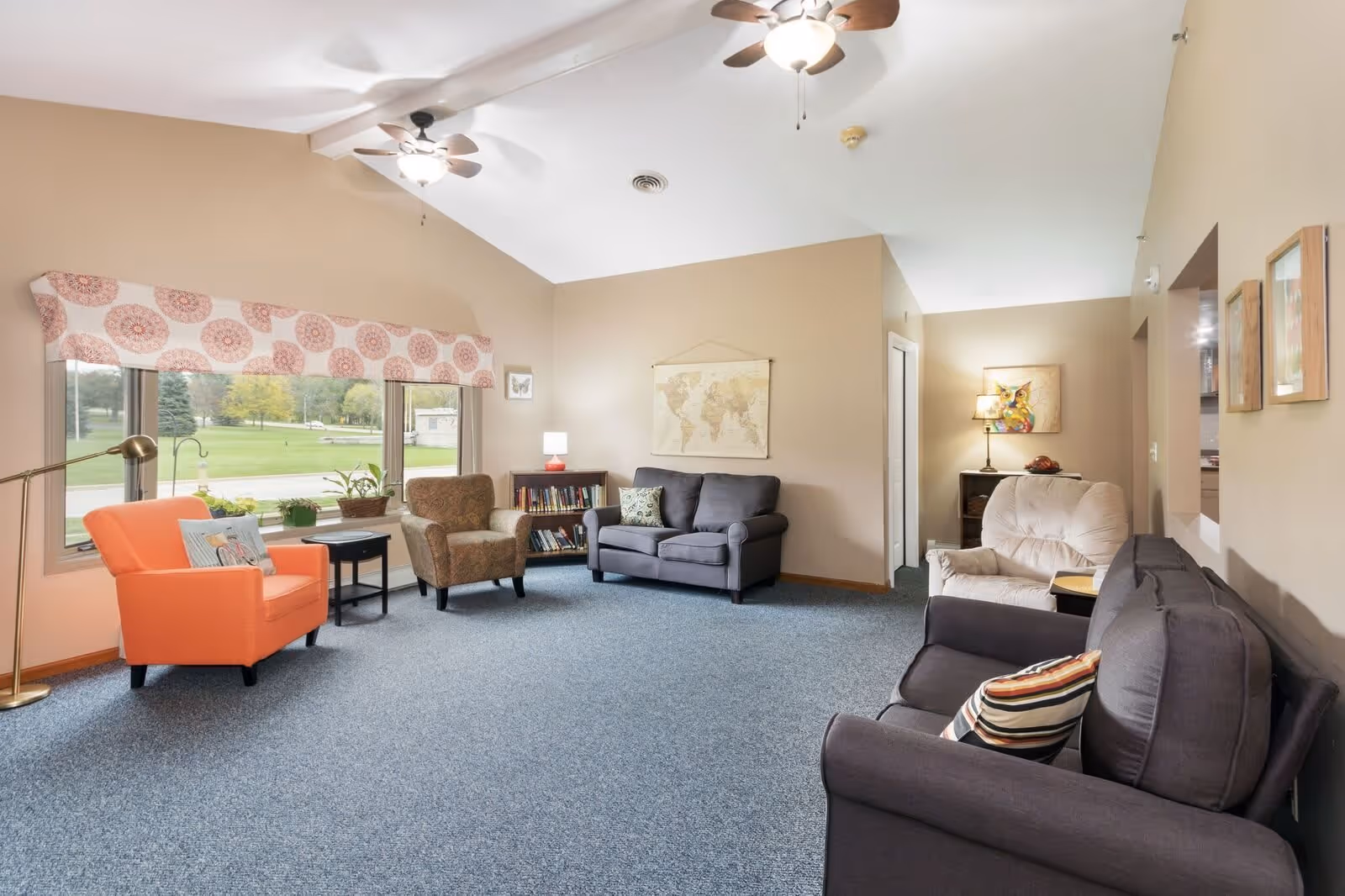 A bright and spacious living room with beige walls and blue carpet. The room features multiple seating options including an orange armchair, a patterned armchair, a gray loveseat, and a beige recliner. There are two ceiling fans with lights, a large window with patterned valance overlooking a green outdoor area, a small bookshelf with books and a lamp, and framed artwork on the walls.
