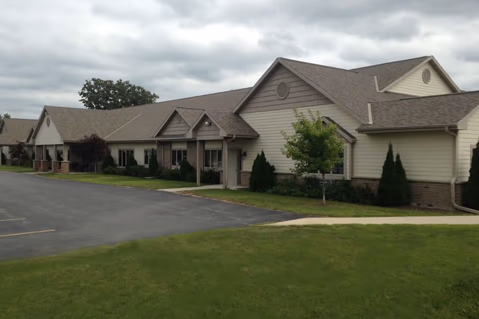 Exterior view of a single-story senior living facility building with beige siding, brown roof shingles, and several windows. The building is surrounded by a well-maintained lawn, small trees, and shrubs. A paved parking lot is visible in the foreground under a cloudy sky.