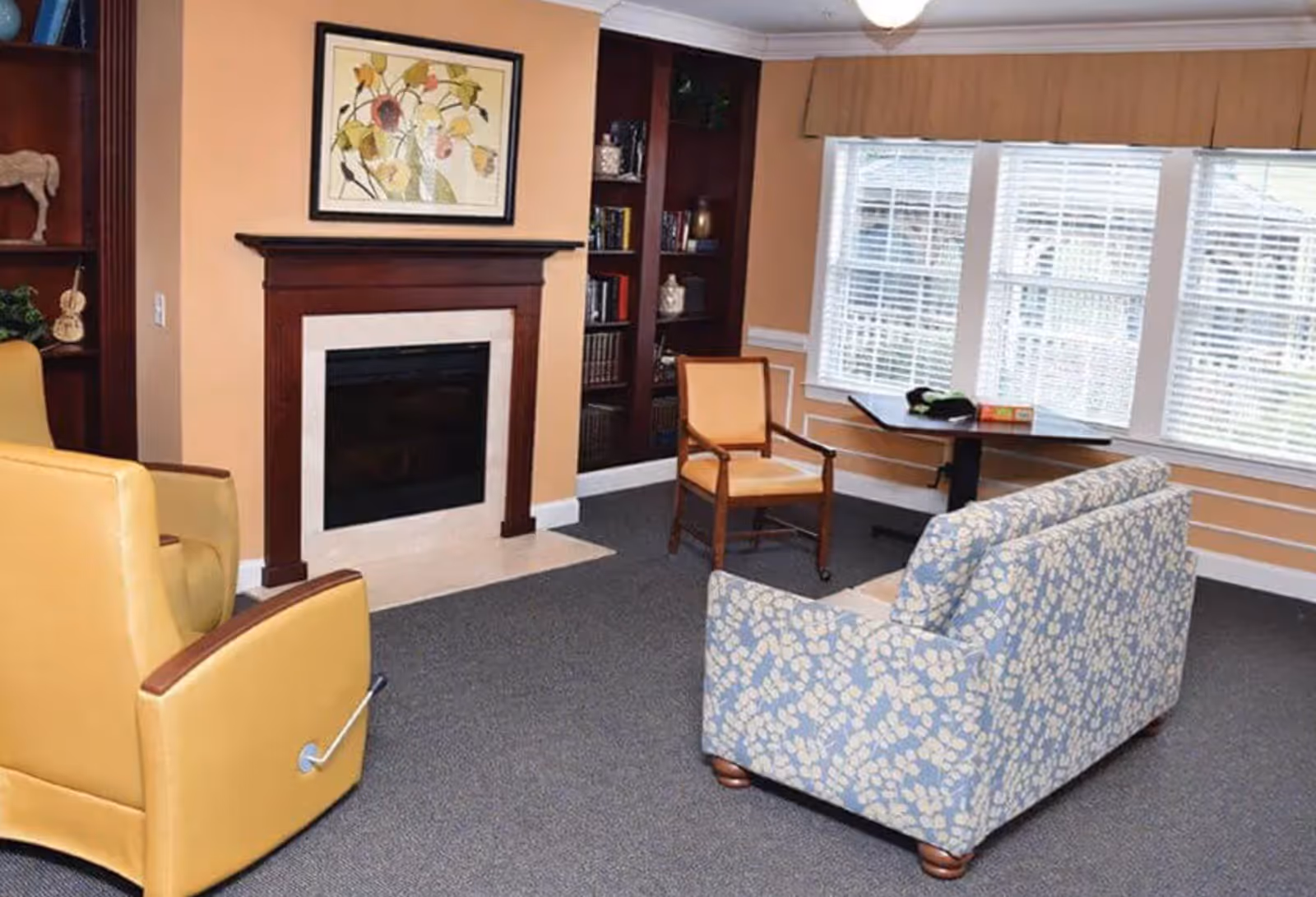 A cozy living room with a fireplace framed by dark wood and beige tiles. There are two yellow armchairs, a wooden chair with a yellow cushion, and a blue patterned sofa. A table is placed near a large window with white blinds, and built-in dark wood shelves flank the fireplace, holding books and decorative items. The walls are painted a warm beige color.