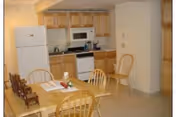 A small kitchen and dining area with wooden cabinets, a white refrigerator, microwave, and stove. There is a wooden dining table with chairs around it, and some items placed on the table.