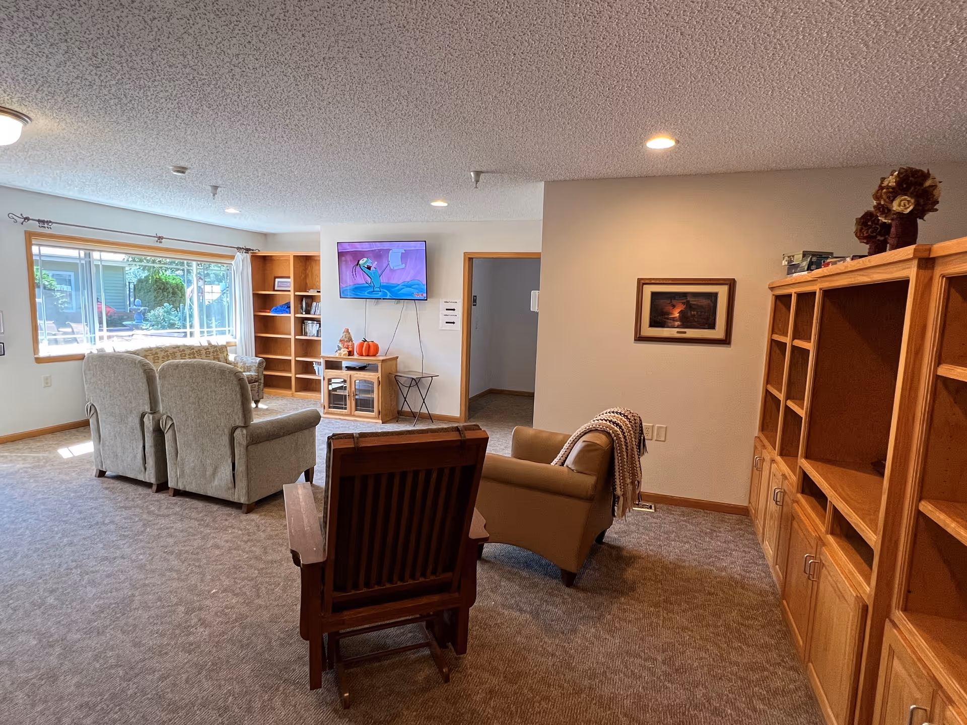 A cozy living room area in a senior living facility with several upholstered chairs arranged facing a wall-mounted TV. There are wooden bookshelves and cabinets along the walls, a large window letting in natural light, and a framed picture on the wall. The carpeted floor and neutral wall colors create a warm atmosphere.