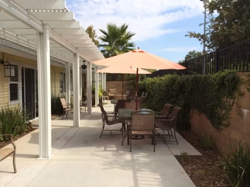 Outdoor patio area with several tables and chairs, an orange umbrella, and a white pergola along the side of a building.
