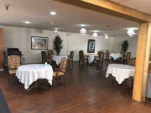 A dining room with several round tables covered with white tablecloths, each surrounded by upholstered chairs. The room has wooden flooring, white walls with framed artwork, and decorative hanging lights. There are also two tall potted plants against the walls.