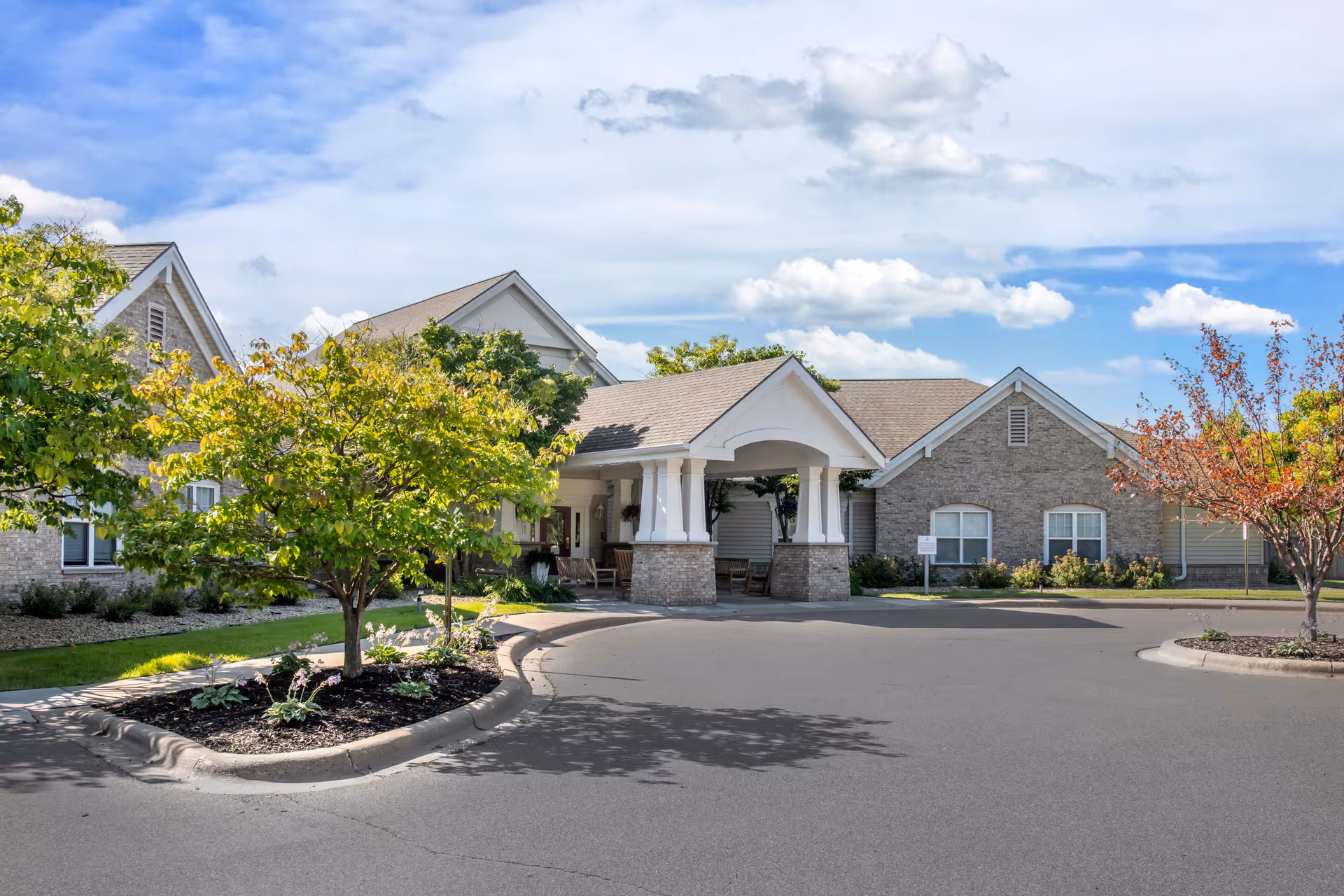 Front entrance of a single-story brick senior living building with a covered porte-cochere, trees, and a circular driveway.