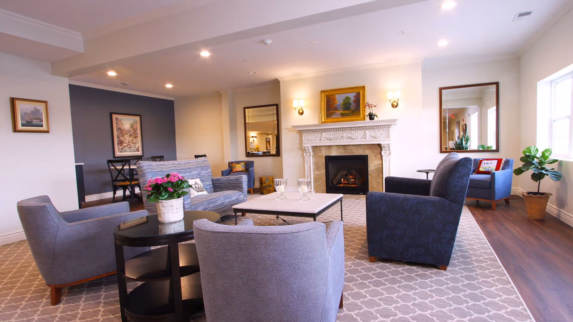 A bright and cozy senior living facility lounge area with comfortable armchairs and a sofa arranged around a coffee table. There is a decorative fireplace with a painting above it, two wall sconces, a large mirror, and a potted plant near a window letting in natural light. The room has a patterned carpet and wooden flooring.
