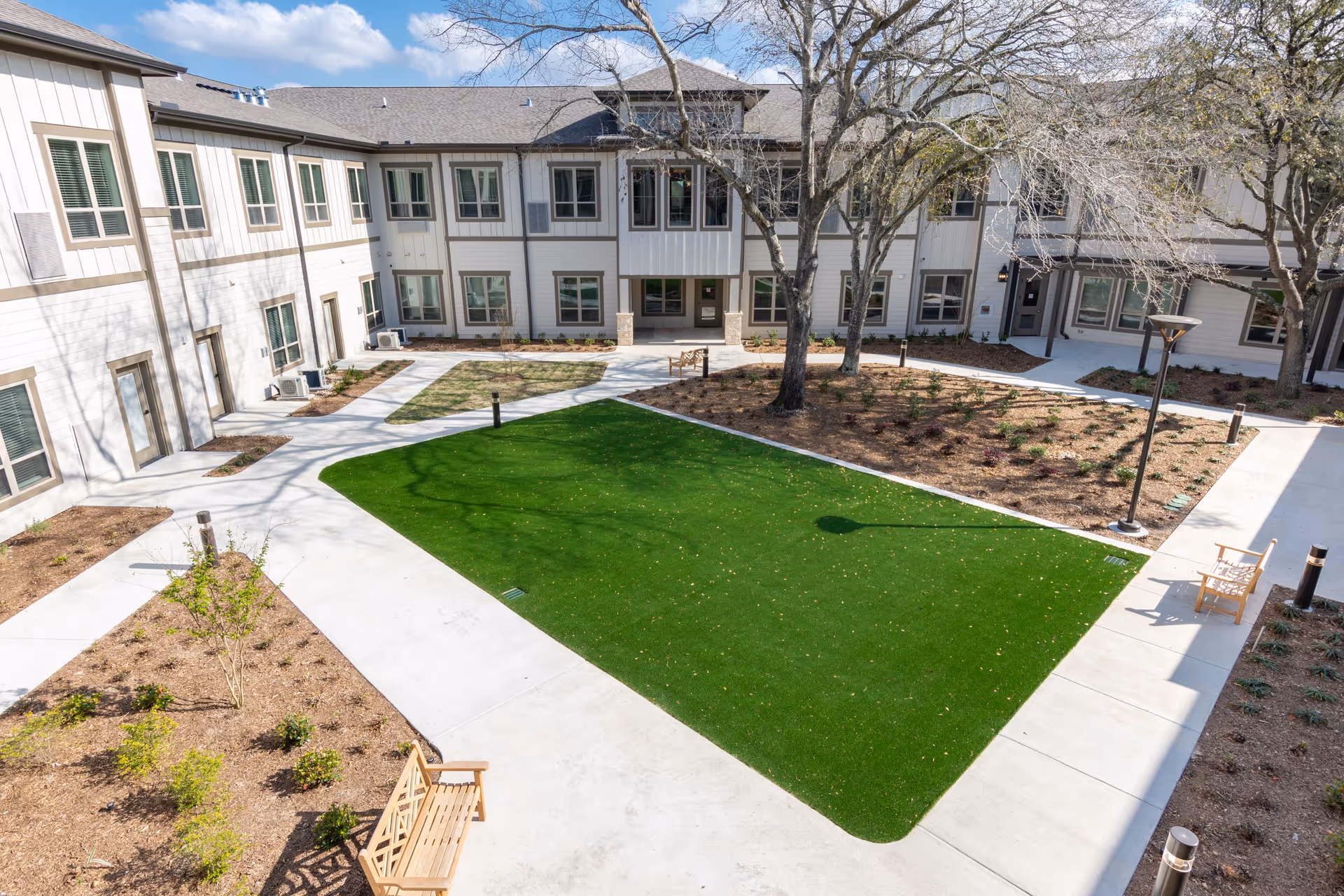 Outdoor courtyard area of StoneCreek of Copperfield Senior Living featuring a well-maintained green lawn, concrete walkways, several wooden benches, leafless trees, and a two-story building with multiple windows surrounding the courtyard.