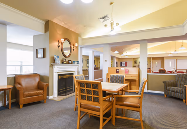 A cozy senior living common area with a wooden table and four chairs in the center, a brown leather armchair to the left, and a fireplace with a round mirror and wall sconces above it. The room has warm lighting, carpeted floors, and additional seating visible in the background.