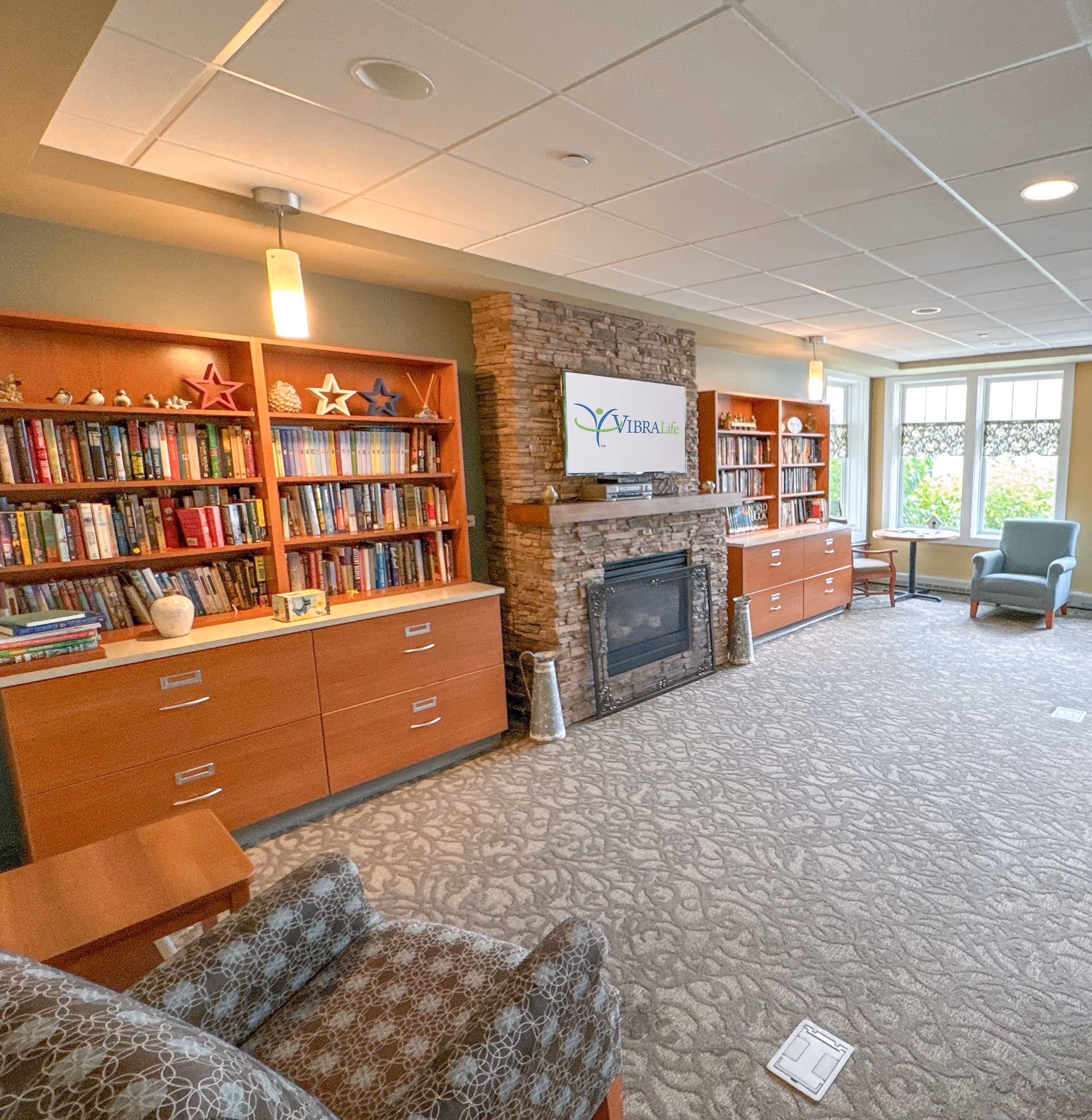 A cozy living room area in a senior living facility with patterned carpet, two wooden bookshelves filled with books and decorative items, a stone fireplace with a mounted TV displaying the VibraLife logo, and comfortable armchairs near large windows letting in natural light.