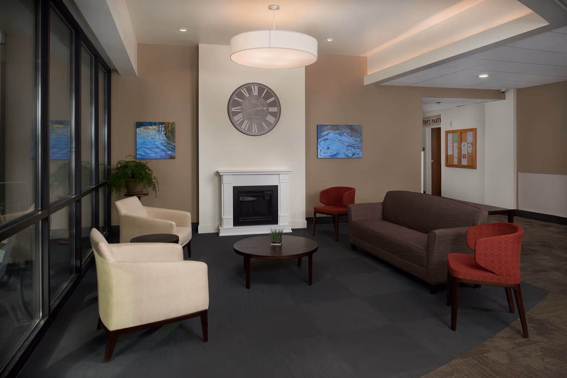 A cozy sitting area in a senior living facility with a brown sofa, two beige armchairs, two red chairs, a round wooden coffee table with a small plant, a white fireplace, a large wall clock above the fireplace, and two paintings on the walls. Large windows line one side of the room, allowing natural light to enter.