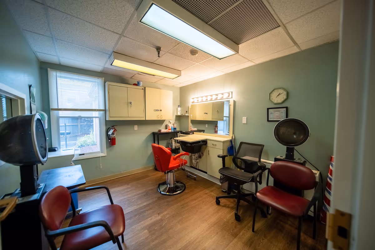 An interior salon room with red styling chairs, a shampoo sink, a hooded dryer, a lighted mirror, and storage cabinets.