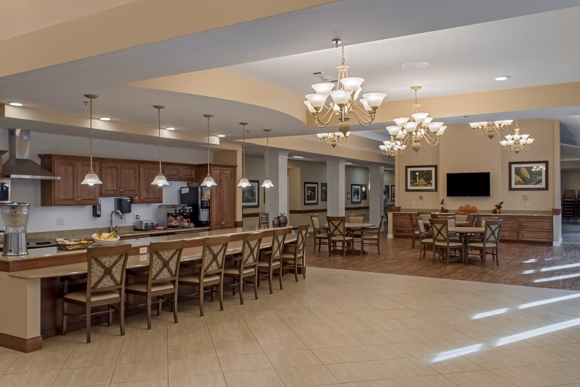 A spacious senior living facility dining area featuring a long counter with multiple chairs, pendant lights hanging above, and a kitchen area with wooden cabinets and appliances in the background. The room also includes several round dining tables with chairs, chandeliers, framed artwork on the walls, and a flat-screen TV mounted on the far wall.