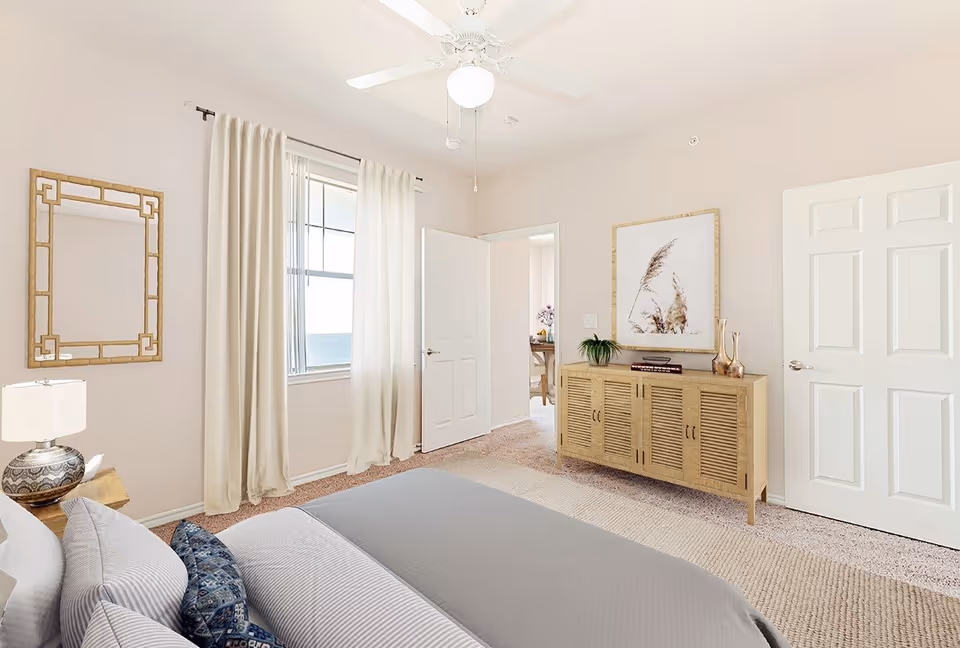 Bright neutral bedroom with a bed in the foreground, a window with curtains, ceiling fan, and a wooden dresser against the wall.
