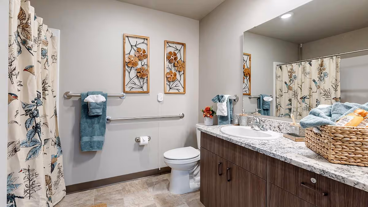A clean and well-decorated bathroom with a toilet, a sink with a granite countertop, a large mirror, and a shower with a floral and bird patterned curtain. There are two framed floral wall decorations above the toilet, teal towels hanging on grab bars, and a wicker basket with toiletries on the counter.