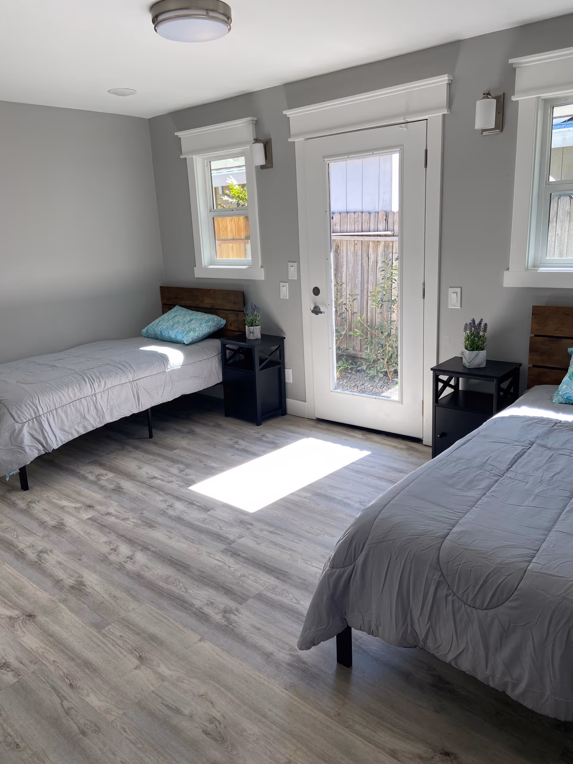 A bright bedroom with two single beds, each with a gray comforter and a teal pillow. Between the beds are two black nightstands, each with a small potted plant. The room has light gray walls, two small windows, and a glass door leading outside. The floor is light wood laminate.