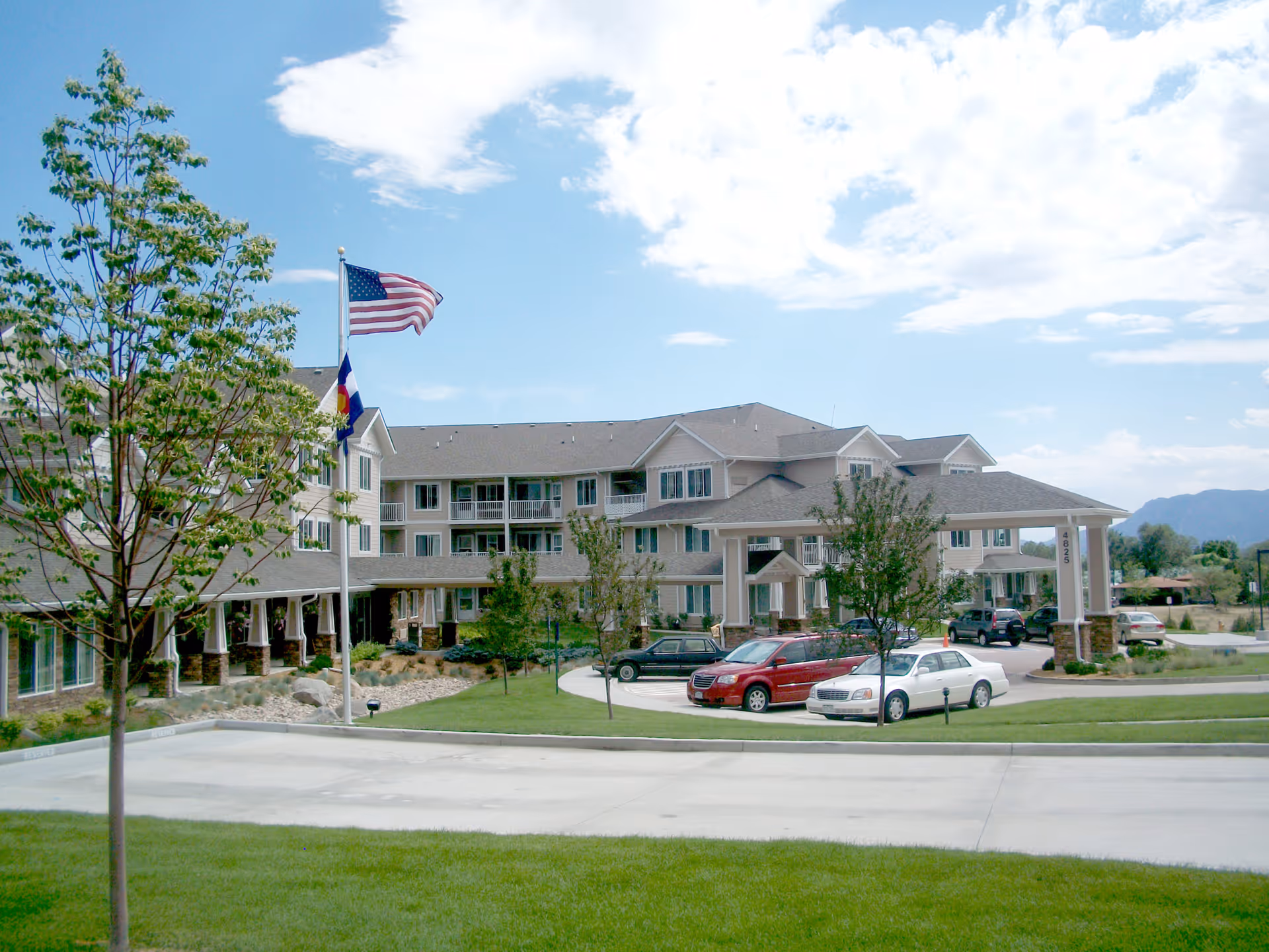 Exterior view of Summit Glen Gracious Retirement Living facility showing a large multi-story building with a covered entrance, several parked cars, green lawn, trees, and two flagpoles with the American and Colorado flags against a partly cloudy sky.