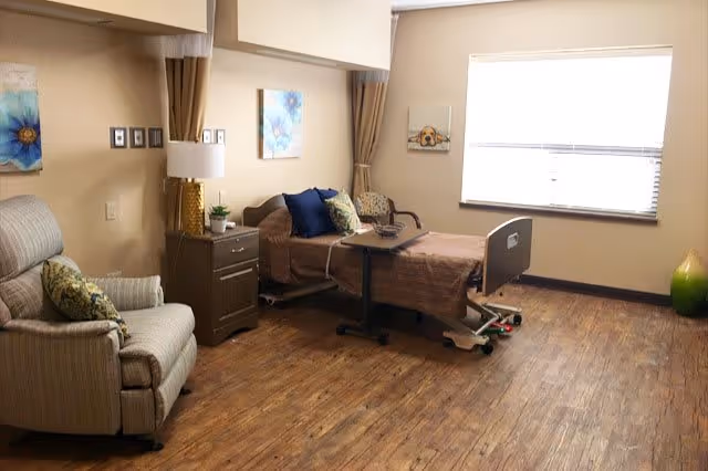 A senior living facility bedroom with a hospital-style bed covered in a brown blanket and several pillows. Next to the bed is a small nightstand with a lamp and a plant. There is a comfortable armchair with a patterned cushion on the left side. The room has wooden flooring, beige walls, a large window with blinds, and decorative wall art.