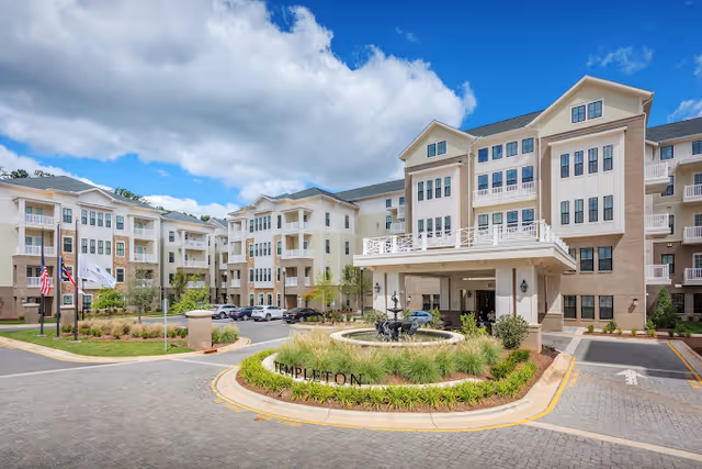 Exterior view of a large multi-story senior living facility with beige and white walls, multiple balconies, and a covered entrance. There is a circular driveway with a landscaped island featuring a fountain and the word 'TEMPLETON' displayed. Flags are visible on the left side, and several cars are parked near the building under a partly cloudy blue sky.
