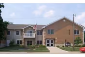 Exterior view of a two-story senior living facility building with a mix of brick and siding. There is a small balcony with an American flag, several windows, and a main entrance with a sidewalk leading up to it. A red car is parked on the right side of the image.