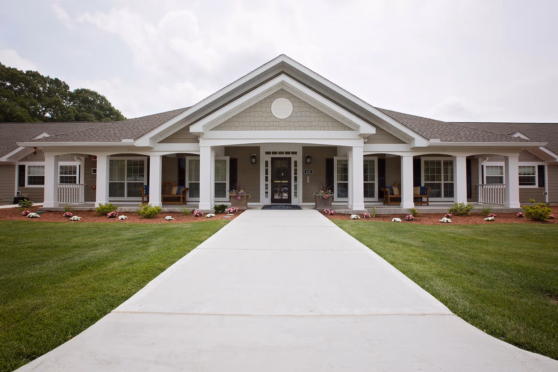 Front exterior view of a single-story senior living facility building with a wide concrete walkway leading to the main entrance. The building features a gabled roof, white columns, multiple windows, and landscaped flower beds along the front.