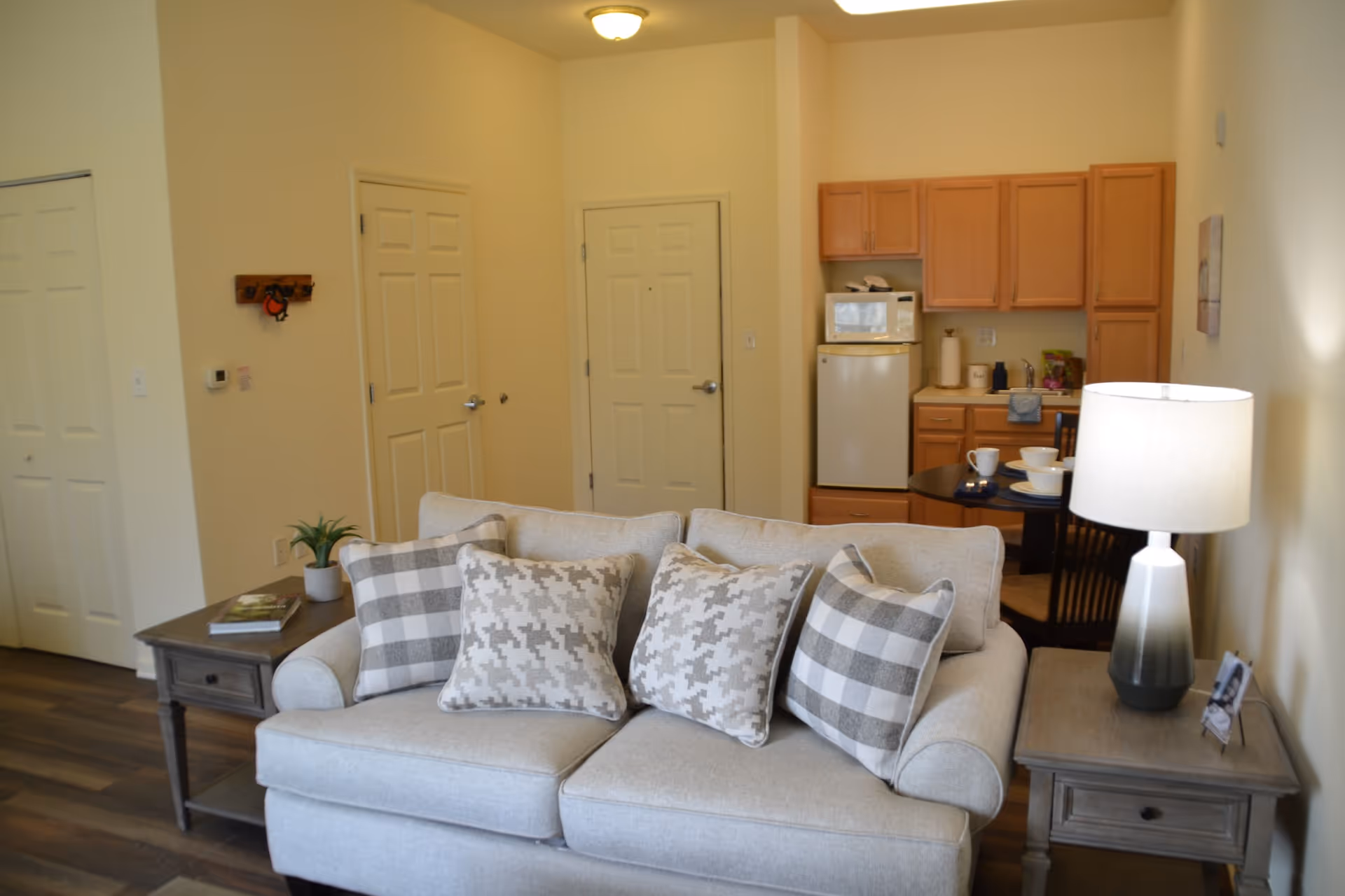 Open-plan living area with a sofa and patterned pillows in the foreground, side tables and a lamp, and a small kitchenette and dining table in the background.