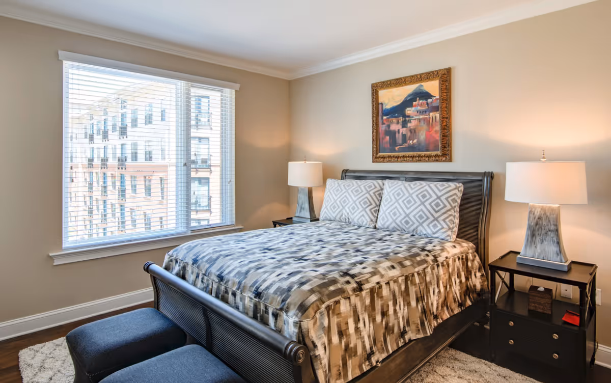 A bright, furnished bedroom featuring a large bed with patterned bedding, two nightstands with lamps, a window overlooking an adjacent building, and framed artwork above the headboard.